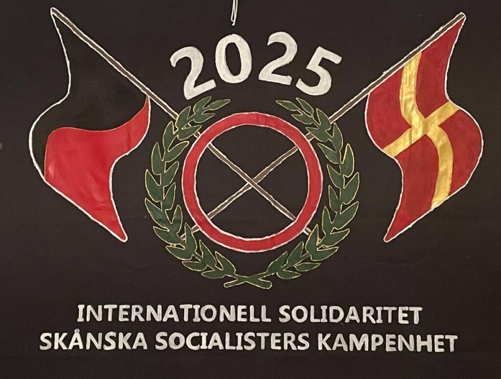 Målad banderoll med SUFs logga. En svartröd och en skånsk flagga korsas i mitten. Text på banderollen "Internationell solidaritet Skånska socialisters kampenhet".